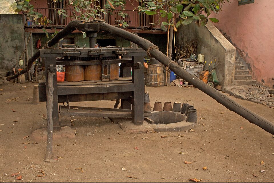 A heavy wooden press with iron rollers and a long draw-beam stands in a dirt yard, with barrels and clay pots around it