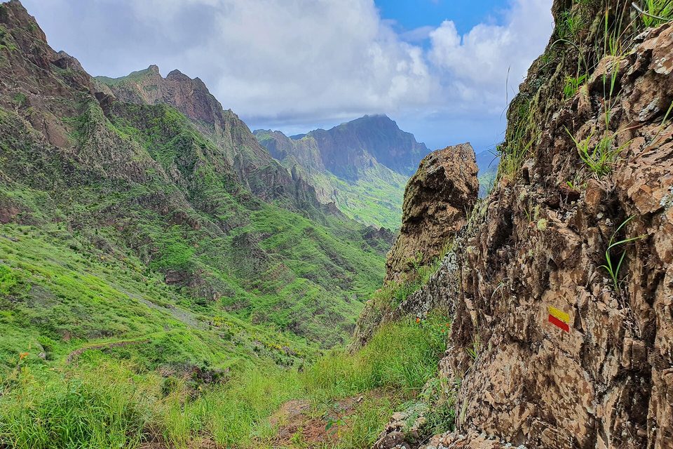 Steep green slopes and ridges under broken cloud, framed by a rocky outcrop with a painted trail mark