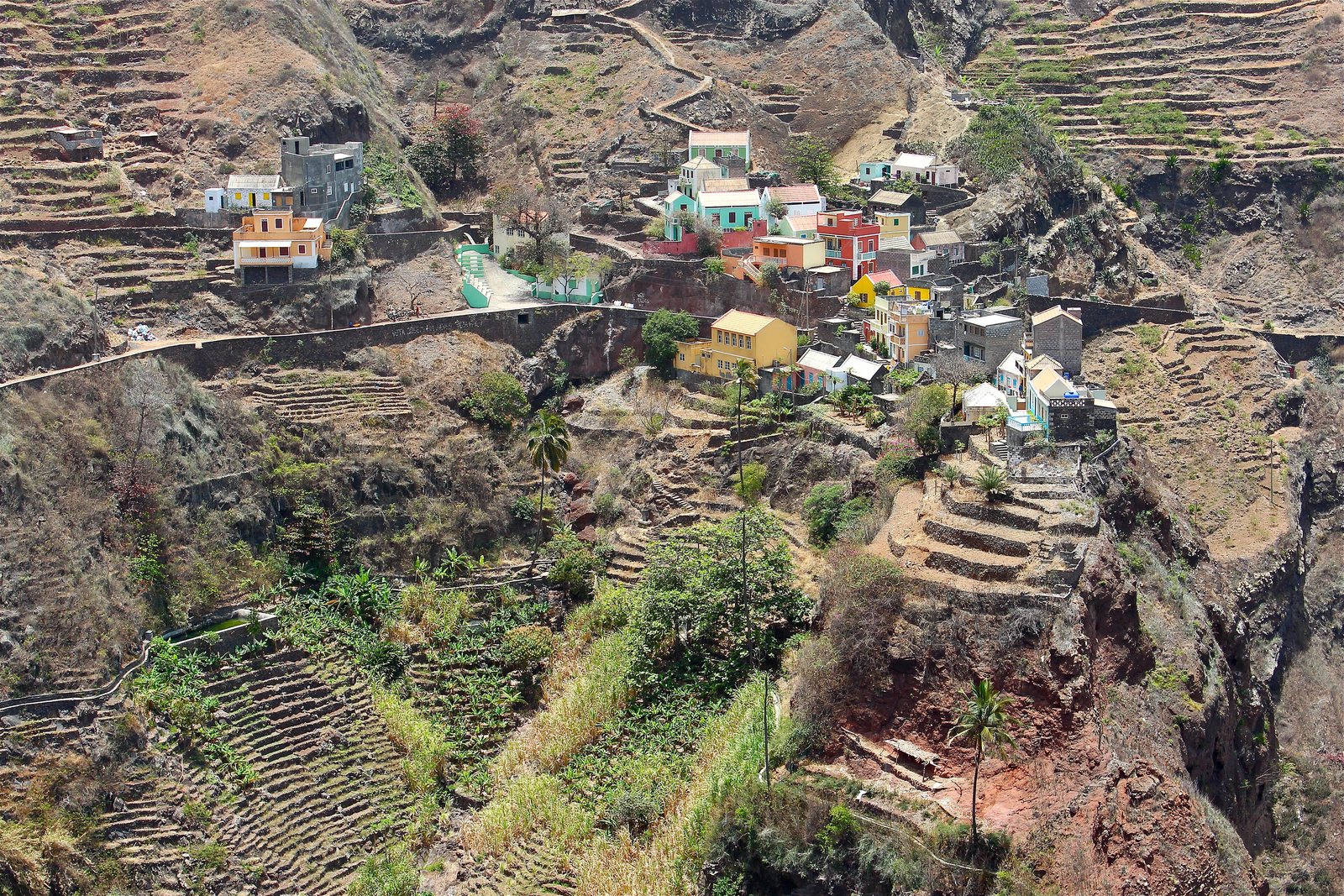 Pink, mint, yellow and red houses cling to a ridge, with stone terraces stepping down into a ravine of banana and cane, and red rock below