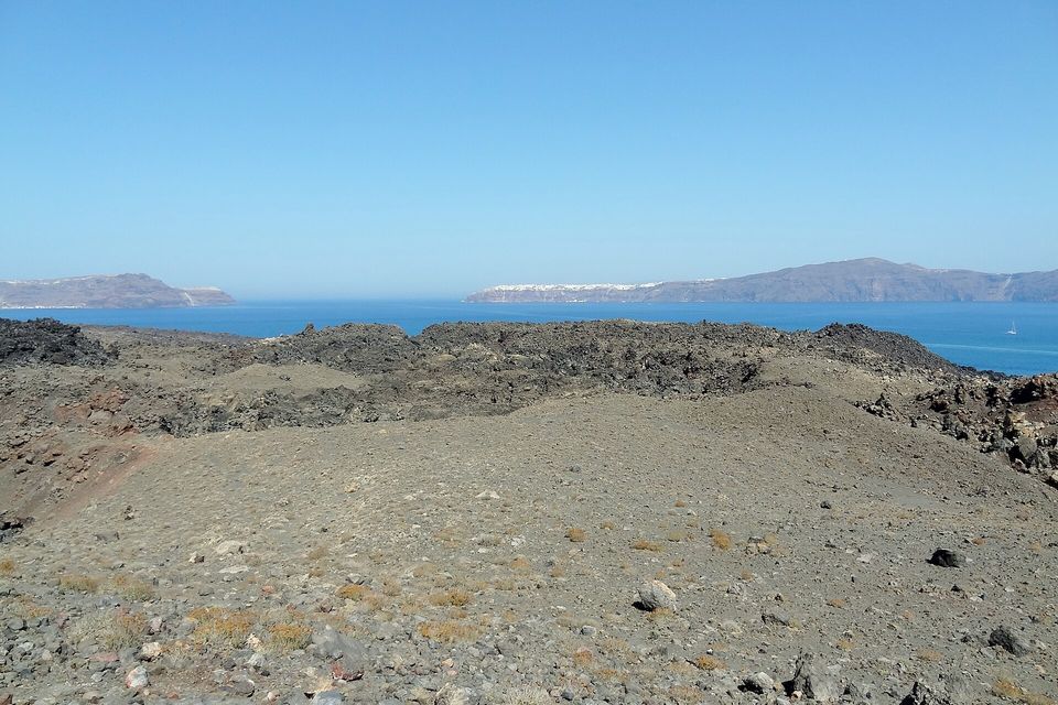 A dark lava field in the foreground, blue sea beyond and two distant islands under a clear sky, one with a white line of buildings along its cliff
