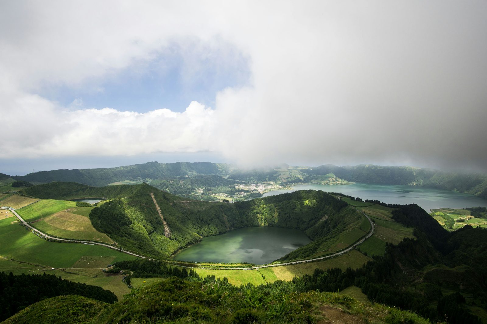 A round crater lake ringed by a road and green pasture, larger lakes and a village beyond under rolling cloud