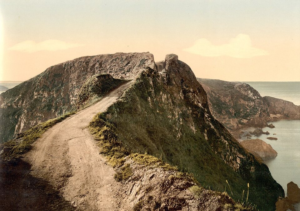 A hand-tinted photochrom of a bare track along a narrow ridge above the sea