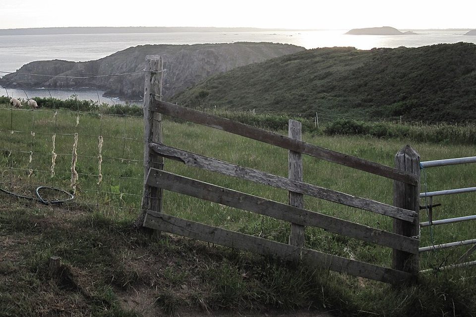 A weathered wooden field gate in a fenced meadow above the sea, with headlands and a distant island in hazy evening light