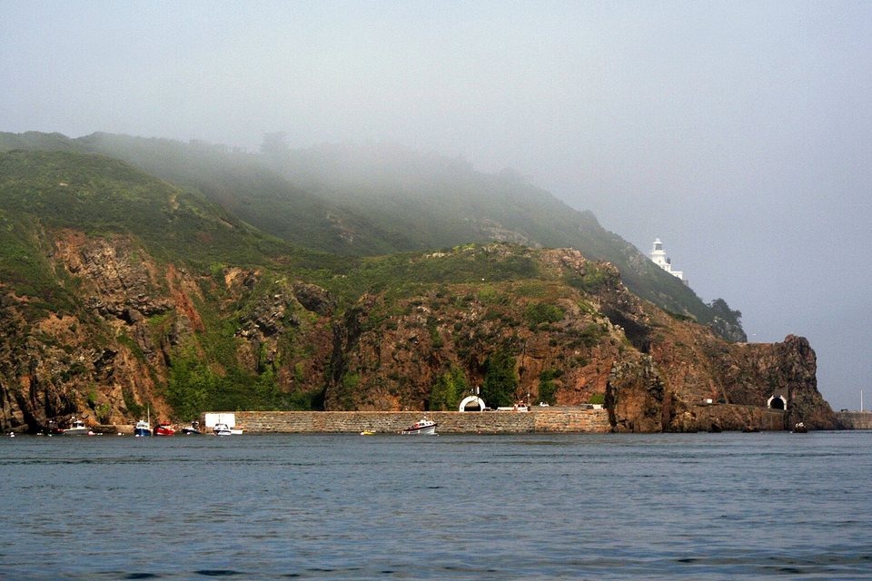 A small stone-walled harbour below red-brown cliffs and green slopes in morning mist, with a white lighthouse on the headland and small boats along the wall