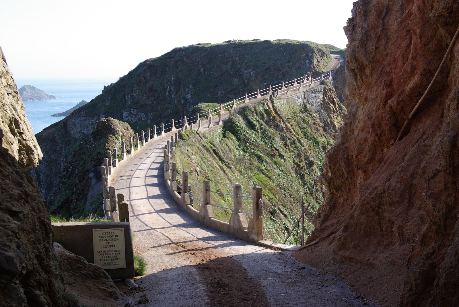 A concrete road with railings running along a narrow ridge between red-brown rock walls, with blue sea at left