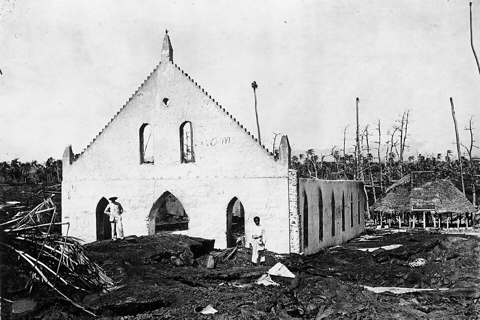 A black-and-white photo of a white church gable with arched openings standing in a field of rough lava, dead palm trunks and a thatched house behind