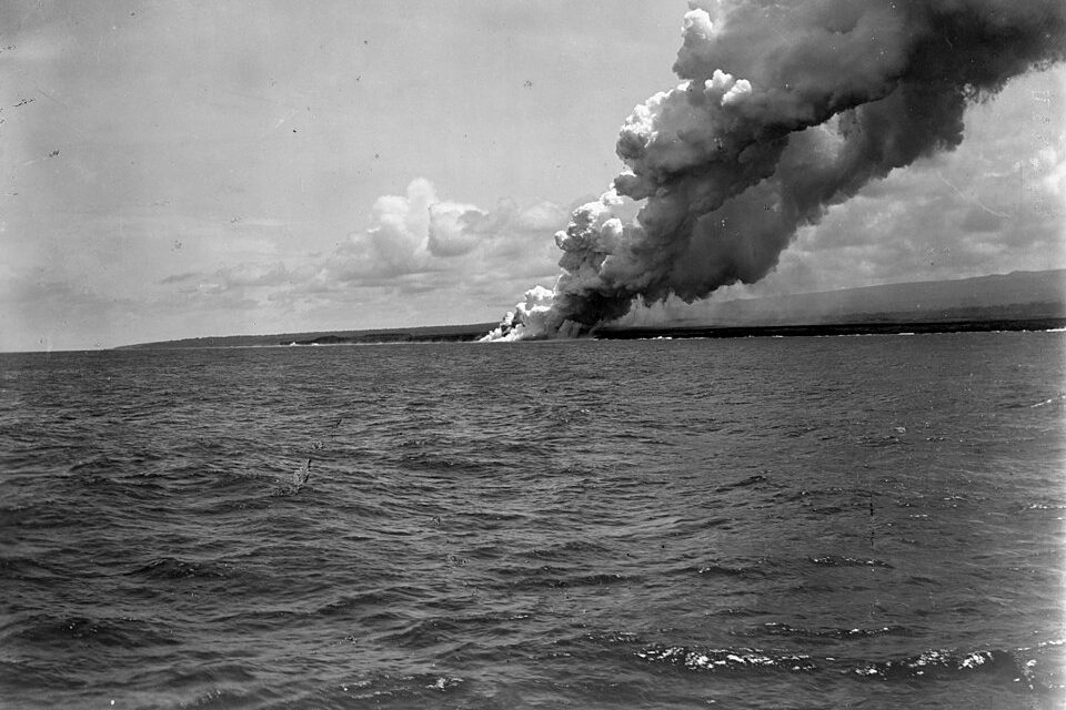 A black-and-white photo of a tall, billowing plume of steam rising from a low coast across a rippled sea