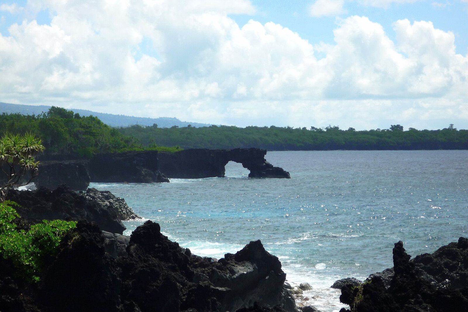 Jagged black lava fills the foreground, and beyond a turquoise bay a natural lava arch stands off a forested shore under bright cumulus