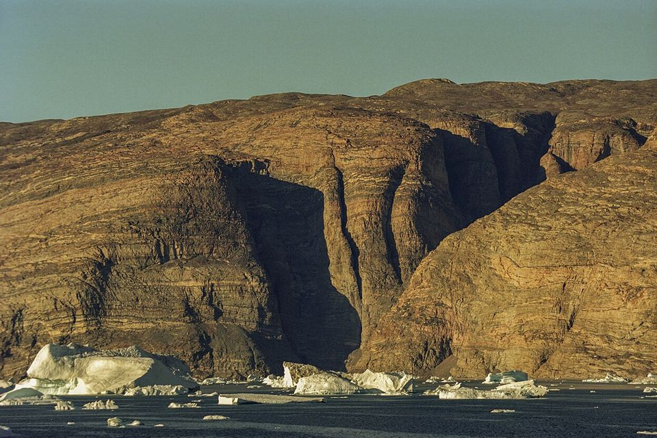 Layered red-brown cliffs cut by deep gullies in low sun, white icebergs on dark water at their foot