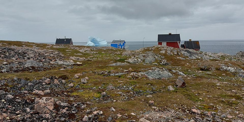 A few red, blue and black wooden houses on bare rocky ground, a large iceberg on grey sea behind