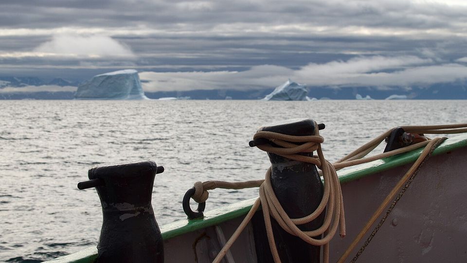 Rope coiled on the black bollards of a ship’s bow, tabular icebergs and low cloud on the water beyond