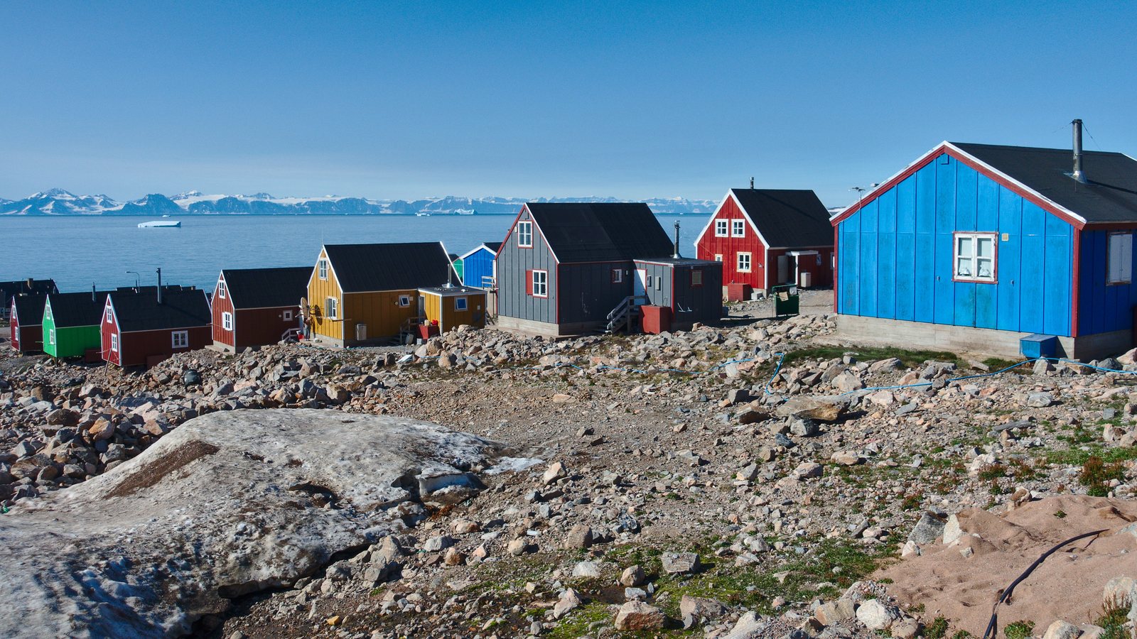 A row of houses painted blue, red, yellow and green standing on bare grey rock and rubble, pale icebergs on the water behind them and snow mountains beyond