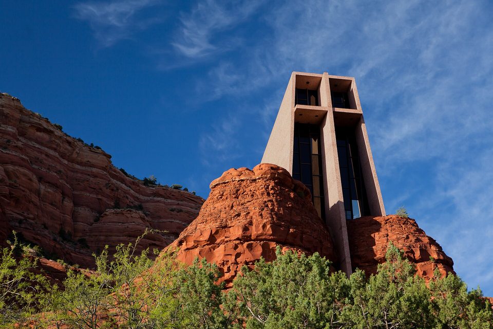 A tall concrete chapel tower with a cross-shaped window frame rising from red sandstone boulders above green trees, deep blue sky