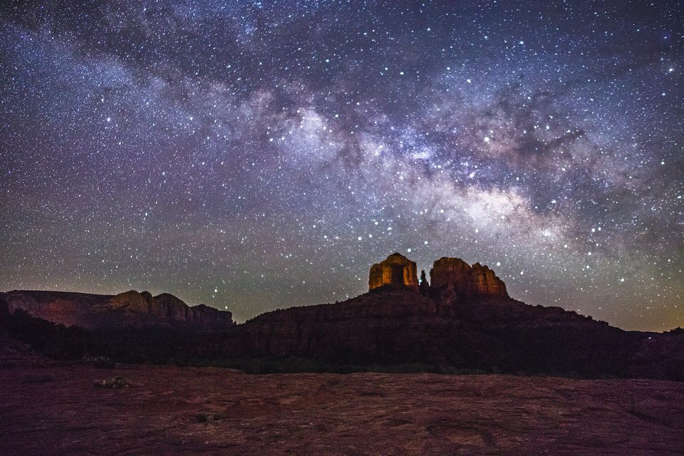 The twin red spires of a sandstone butte faintly lit at night beneath a dense band of stars
