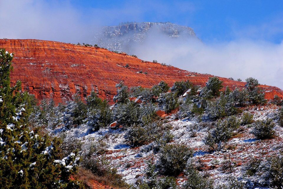 Bright red layered cliffs above juniper slopes dusted with fresh snow, a pale summit half-hidden in low cloud
