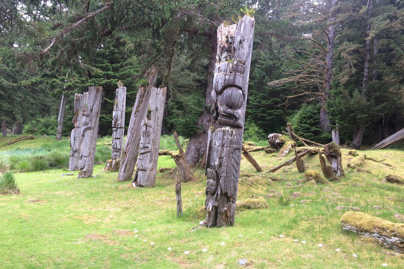 Four grey weathered mortuary poles leaning at different angles on a mossy clearing, carved faces still legible, collapsed longhouse beams beside them and dark spruce forest behind