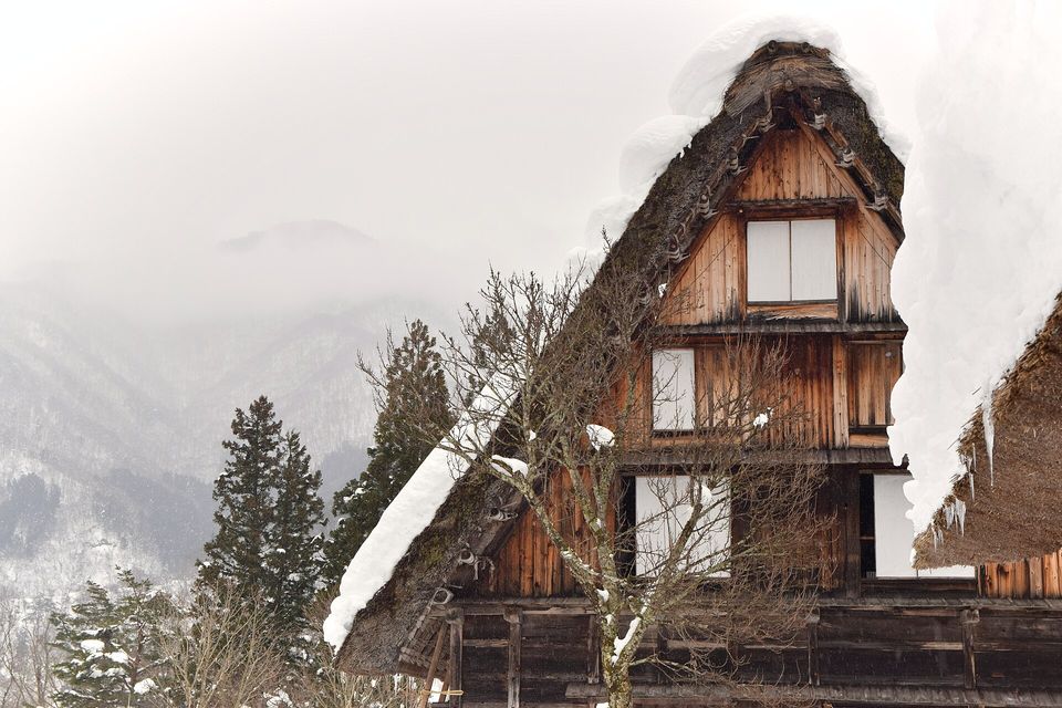 The tall wooden gable of a thatched farmhouse, its roof heavy with snow, rises beside a bare tree against a misty, snow-dusted mountainside