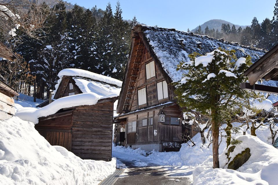 A cleared lane runs between metre-high snowbanks toward a thatched farmhouse with a snow-laden pine, wooden sheds at left, under a clear blue sky