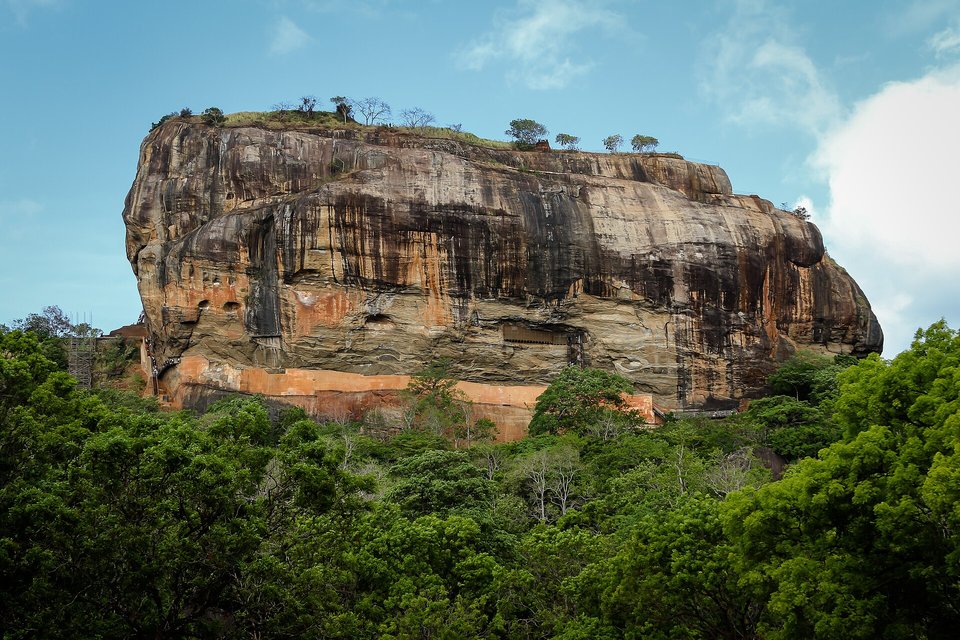 A massive orange, black and grey streaked rock with a flat top crowned with small trees, rising above green forest under a blue sky