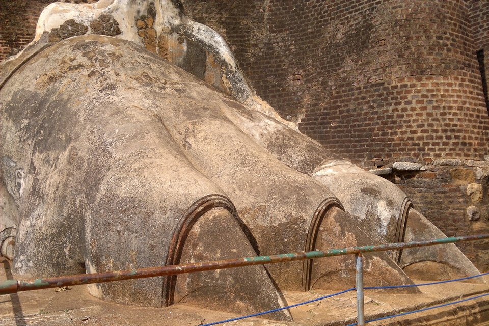 A huge weathered stone paw with three arched claws, a rusted railing and rope in front of it and a brick wall behind
