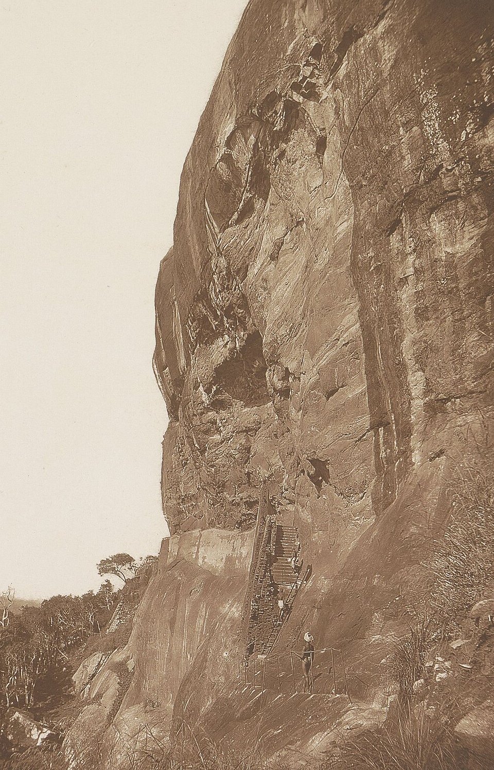 A sepia photograph of a sheer rock wall, a stairway climbing its face to a ledge, a small figure at its foot and scrub below