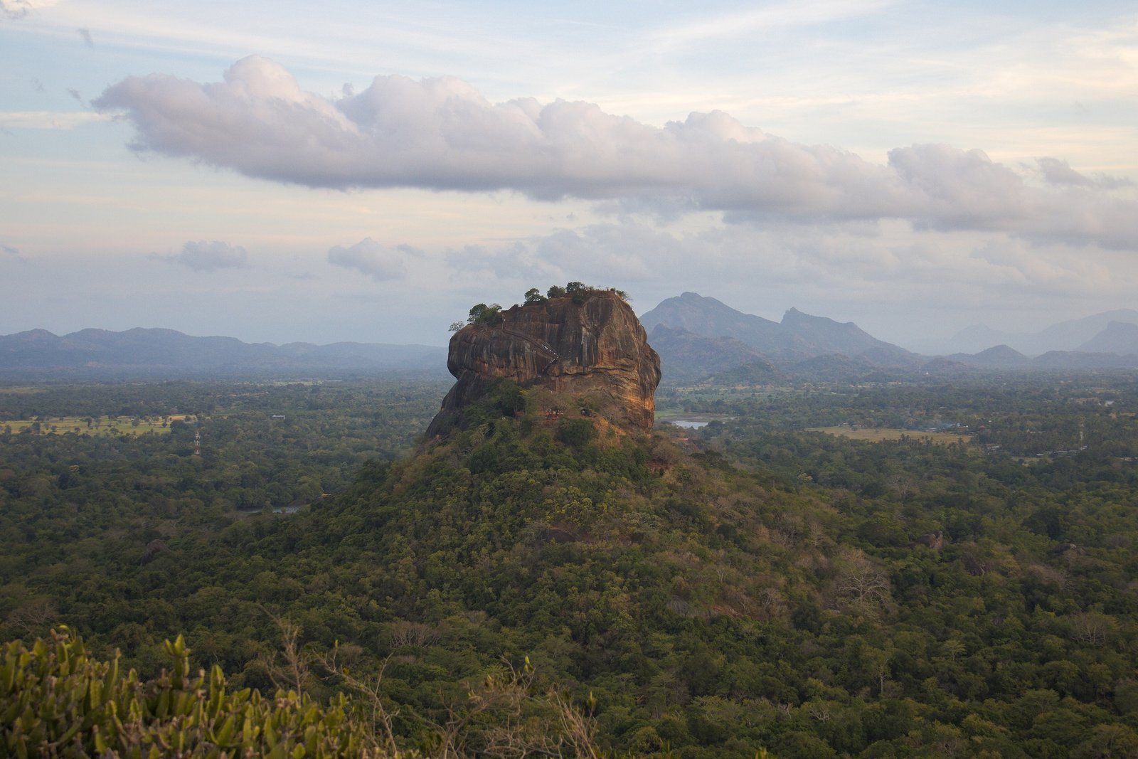 A tall flat-topped rock of orange-brown granite standing alone above dense green forest, low blue hills and a broad cloud beyond