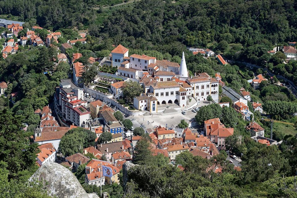 Red-tiled roofs of a hill town around a white palace with a tall white conical chimney, dense forest behind