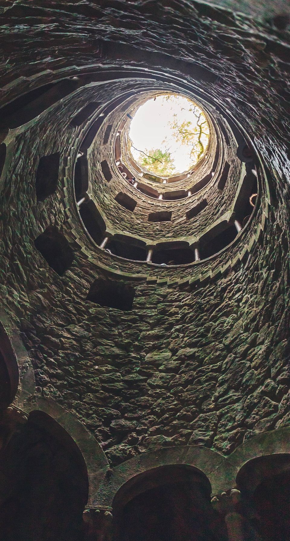 A stone spiral shaft seen from the bottom, ringed by galleries and arches, rising to a disc of daylight and leaves