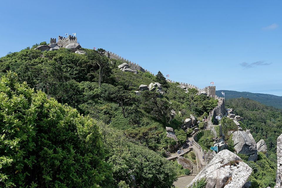 A medieval stone wall and towers running along a boulder-strewn forested ridge under a blue sky, the sea faint at the right