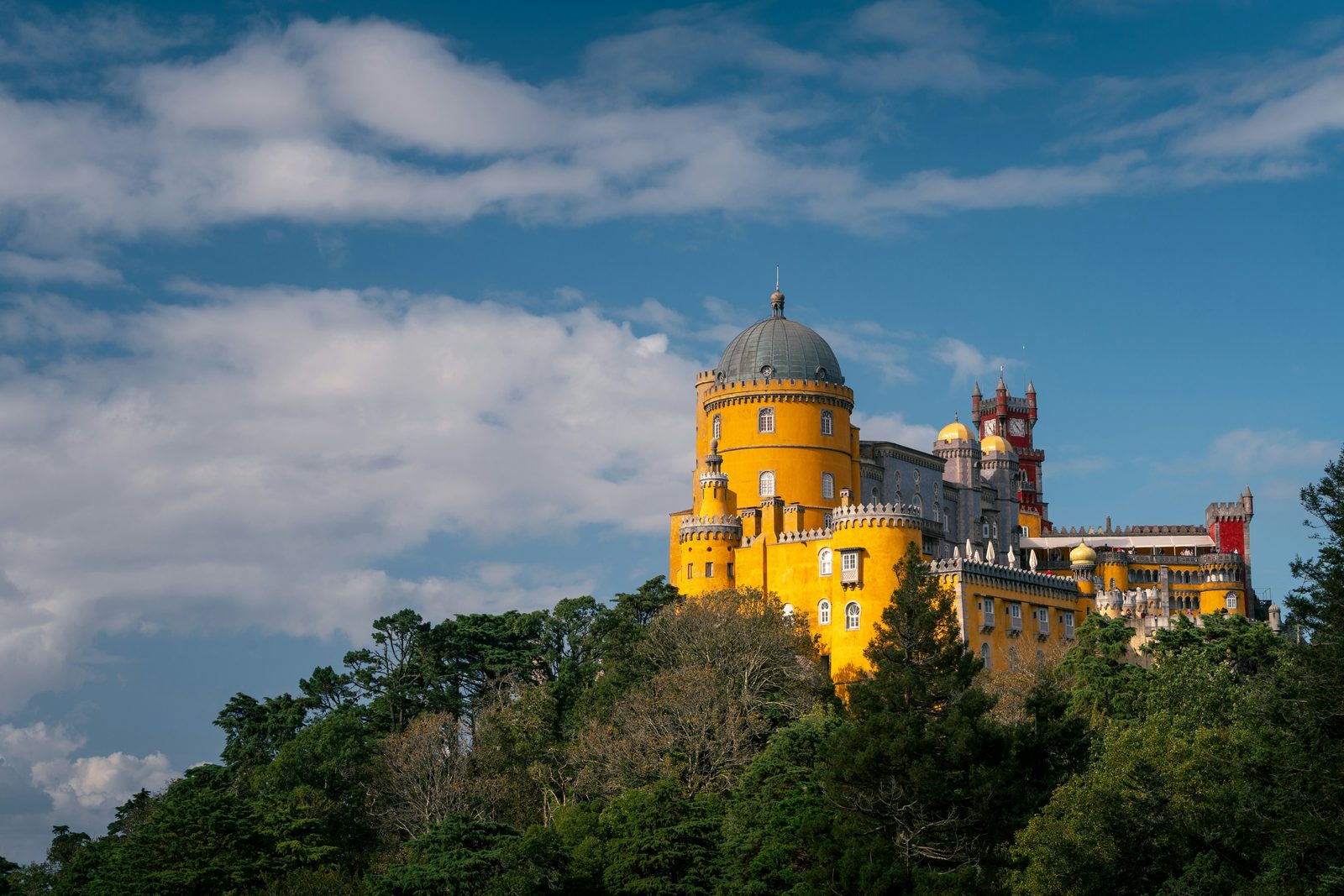 A yellow and red palace on a forested hilltop in low directional light under broken cloud