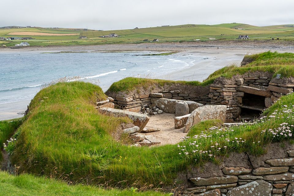 Grass-topped stone walls of a sunken house beside a curving sand beach, sea pinks in the foreground