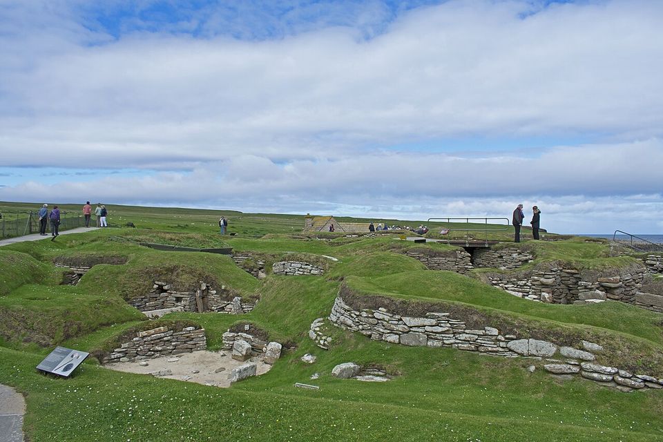 Grassy mounds and sunken stone house walls under a wide sky, a few small figures on a path at the far edge