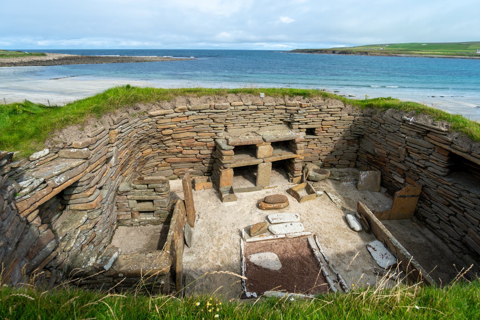 A Neolithic stone house seen from above, walls enclosing a square hearth, stone box beds either side and a two-shelf stone dresser against the far wall