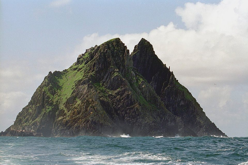 A steep, twin-peaked rock island with green ledges rises straight out of a choppy grey-blue sea