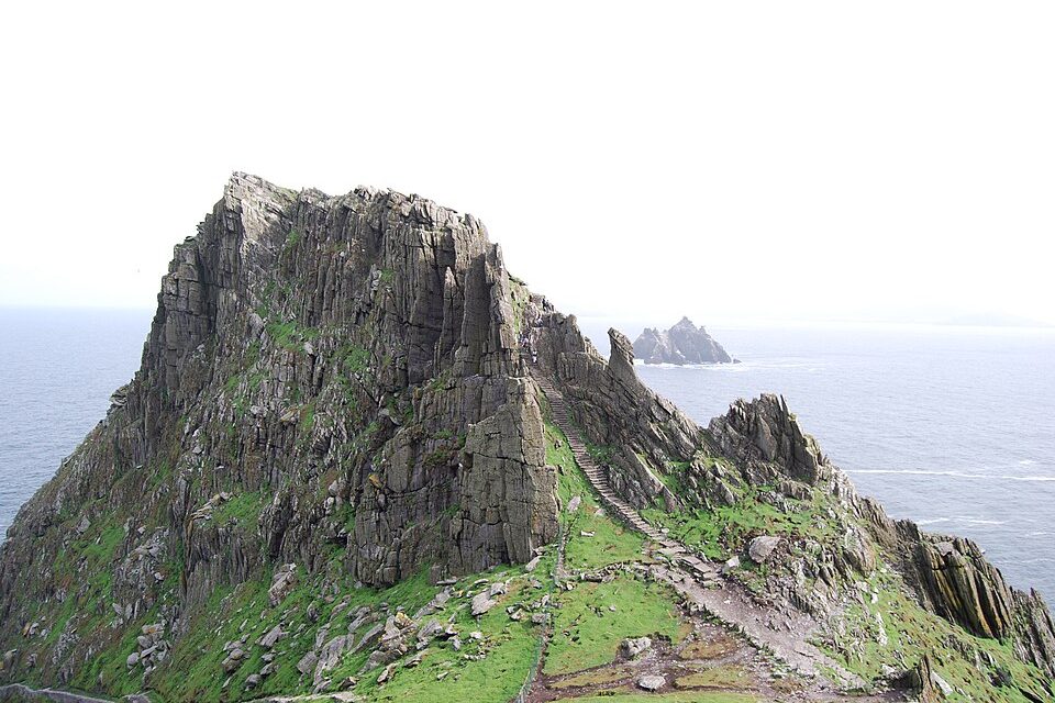 A stone path winds up a green saddle below a tall grey crag, with a small rocky island far out at sea