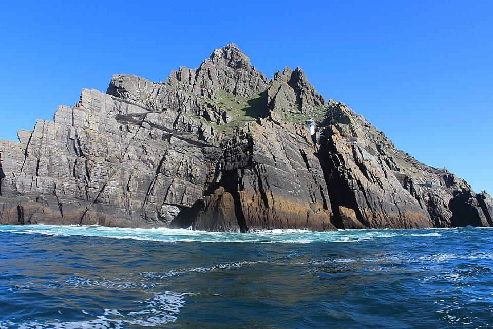 Layered grey cliffs streaked with rust rise from swirling blue water to a jagged summit, a small white lighthouse halfway up
