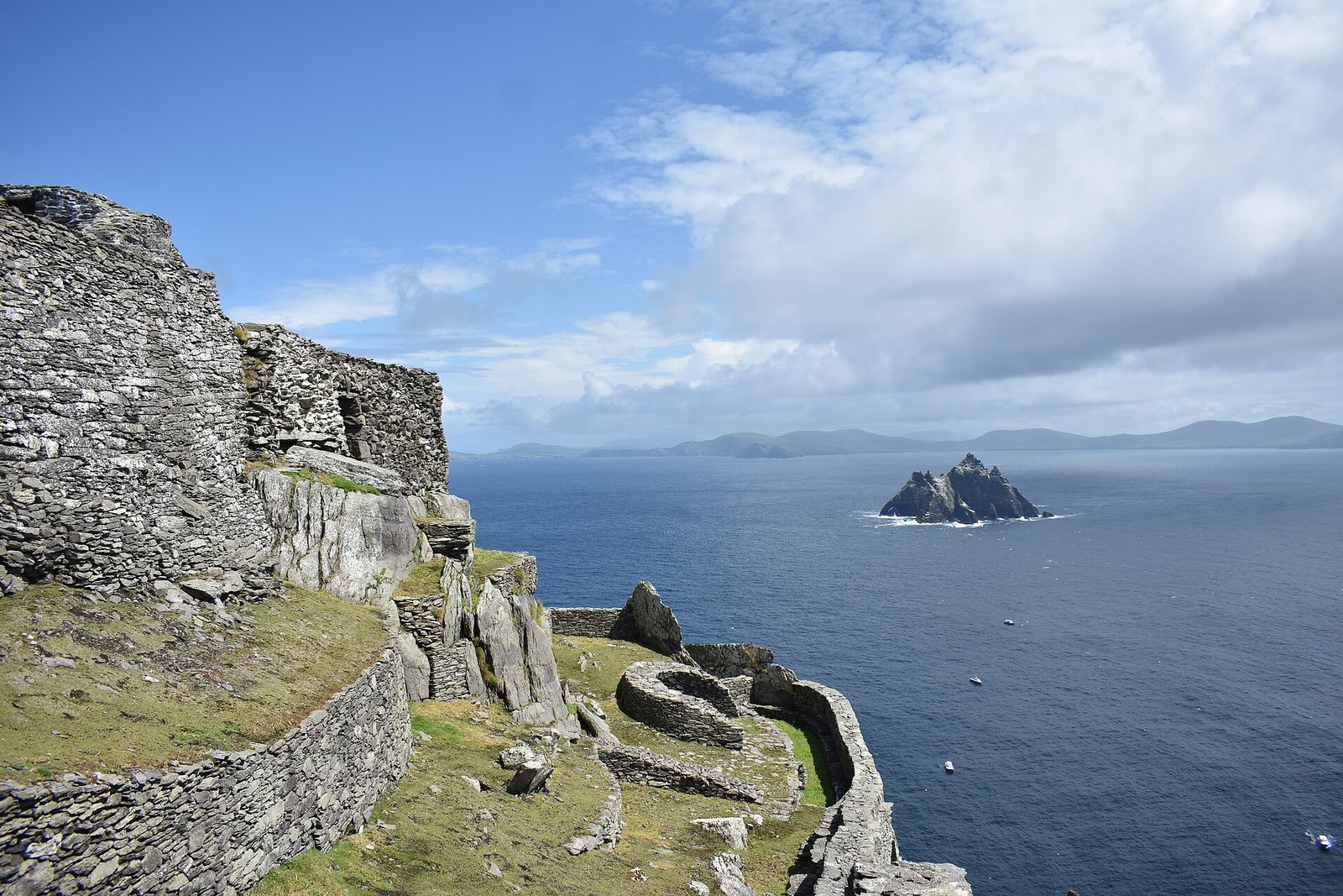 Dry-stone beehive huts and terrace walls stand on a ledge at left, with deep-blue sea, a rocky islet and distant hills under broken cloud at right