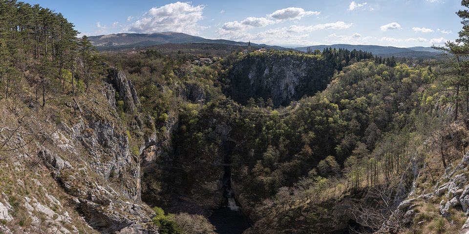 Wooded collapse hollows and a gorge below a village on a cliff, under spring cloud