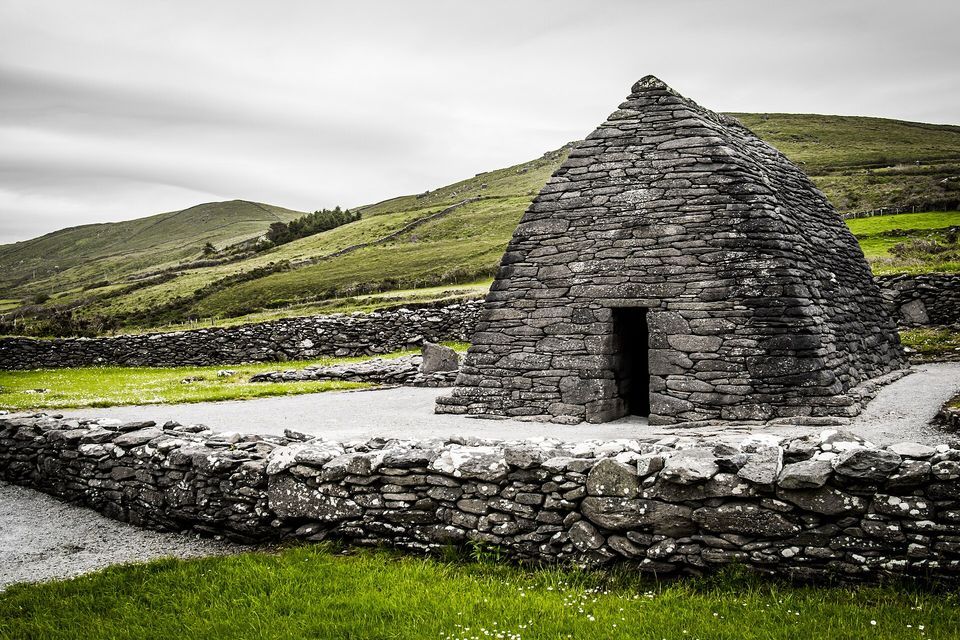 A small boat-shaped church of closely fitted grey stones with a dark doorway, inside a low stone enclosure, green hills under a grey sky