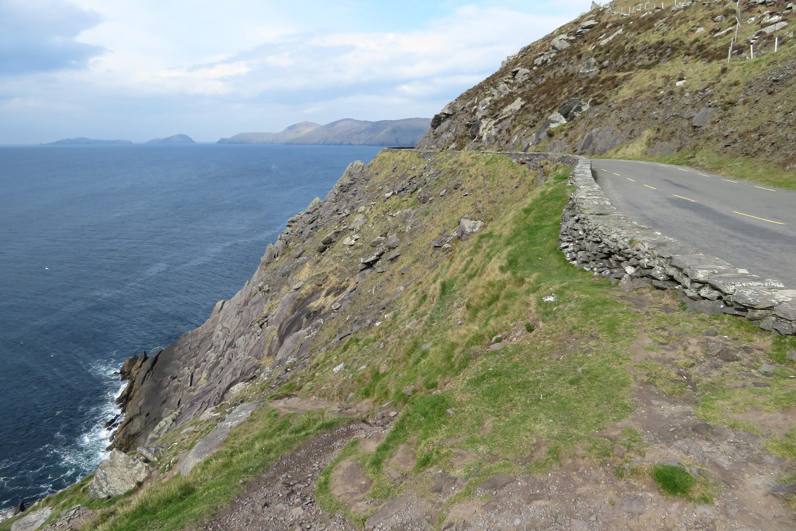 The narrow Slea Head road edged by a dry-stone wall, bending round a steep green headland above a blue sea, the Blasket Islands on the horizon