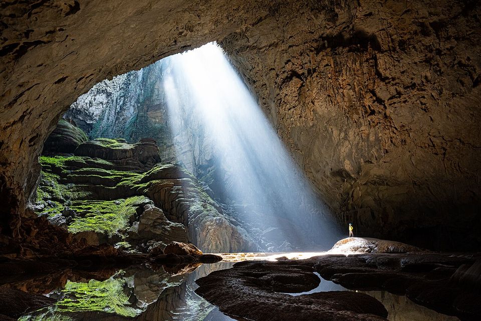 A shaft of sunlight falling into a vast cave chamber onto mossy rock, a tiny figure standing in the light