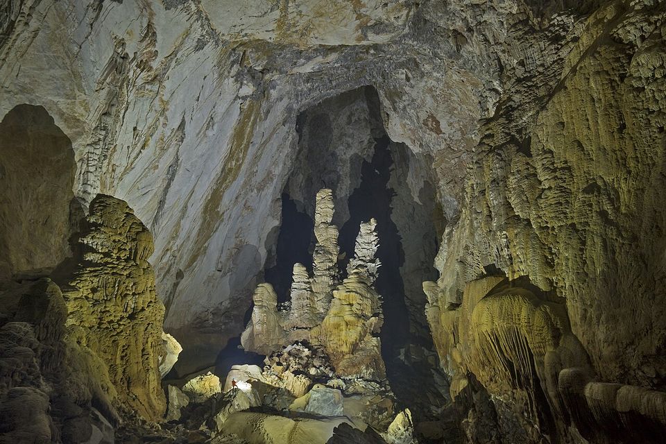 Pale stalagmites like stacked towers rising from a boulder floor in a huge dark passage