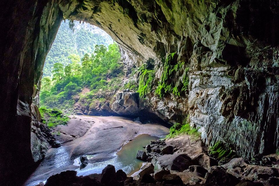 A huge cave mouth framing green jungle, a shallow river running over a sandy floor