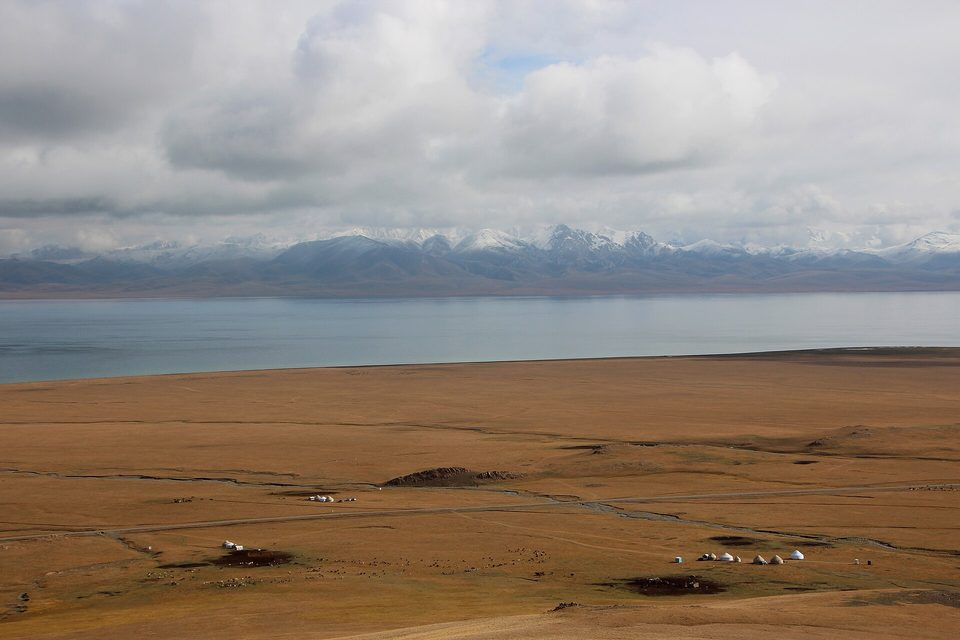 A wide grassy steppe with a blue lake and a range of mountains beyond, a few small yurts scattered across the slope