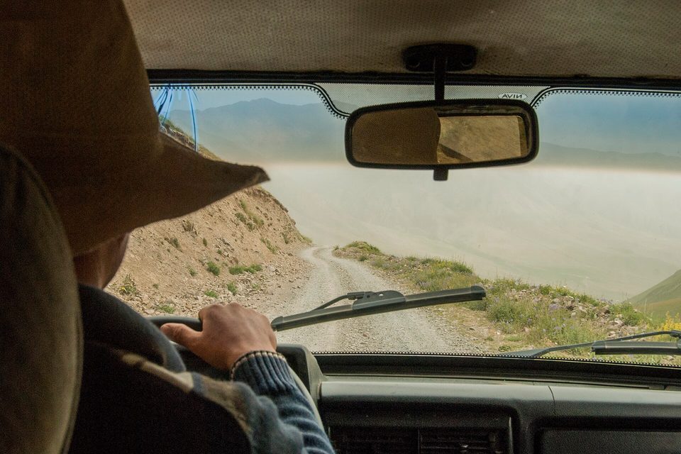 A gravel mountain road dropping away through a car windscreen, the driver seen from behind