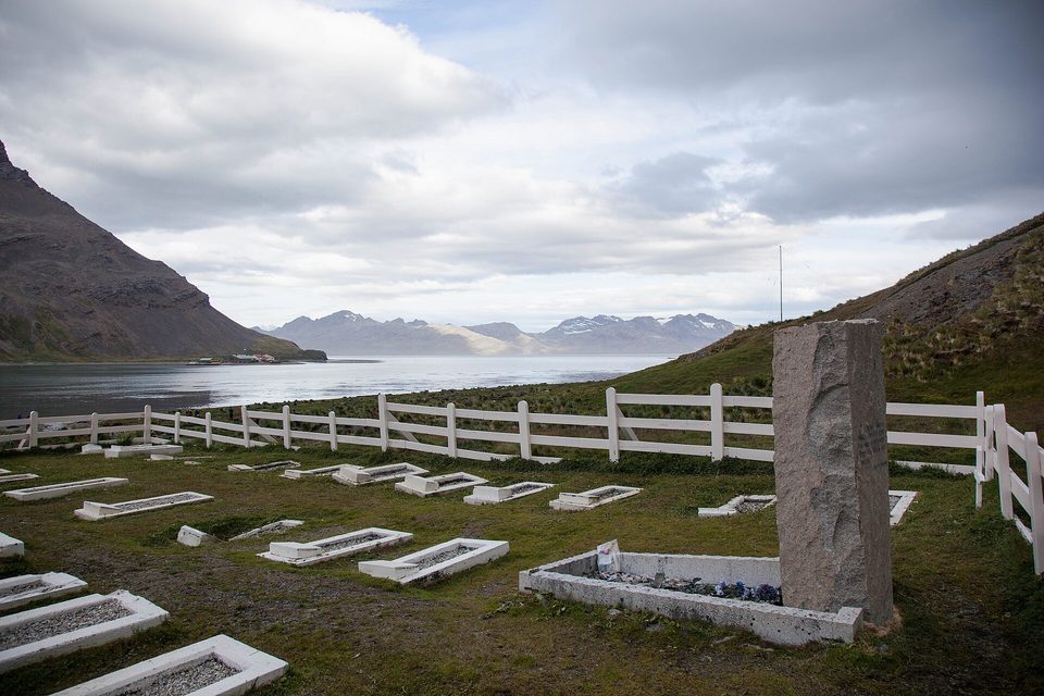 A white-fenced cemetery with a granite headstone, a calm bay and hills behind, no visitors in view