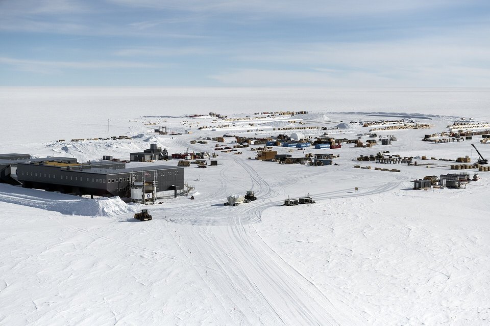 A long dark elevated building at left and a scatter of small camp buildings, vehicles and tracked roads on a flat white plain under a pale blue sky