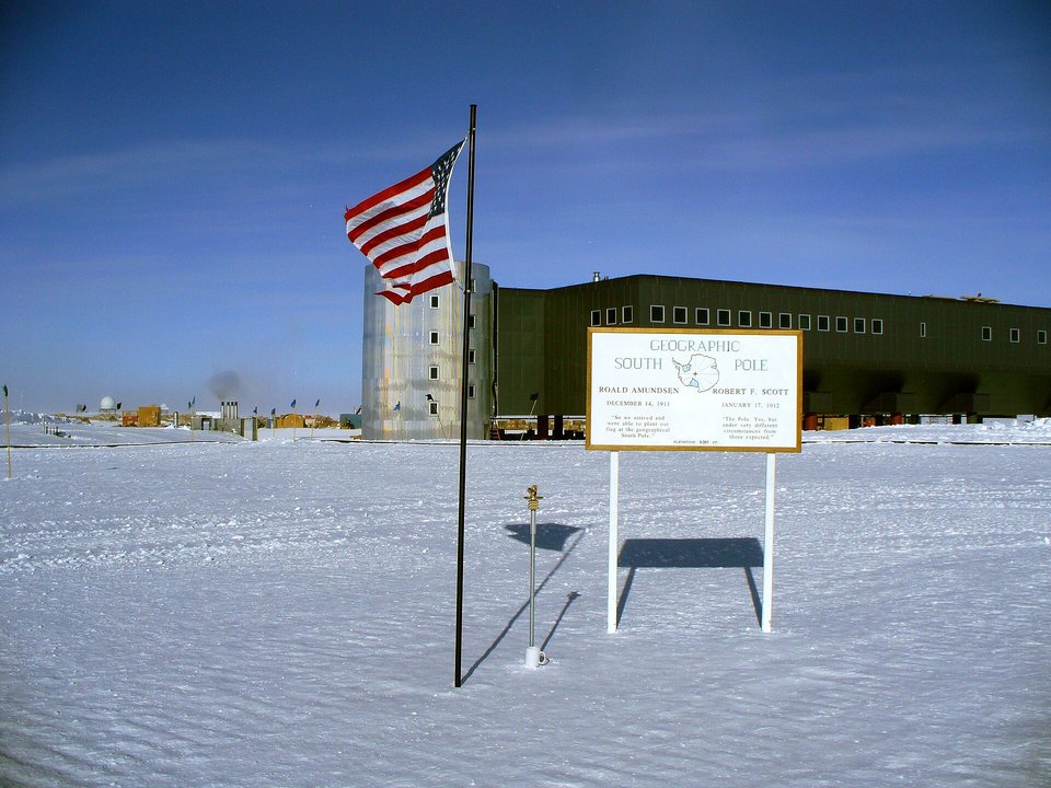 A flag on a tall pole and a white sign reading Geographic South Pole beside a small marker on a post in the snow, a long green elevated building and a silver tower behind, under a deep blue sky