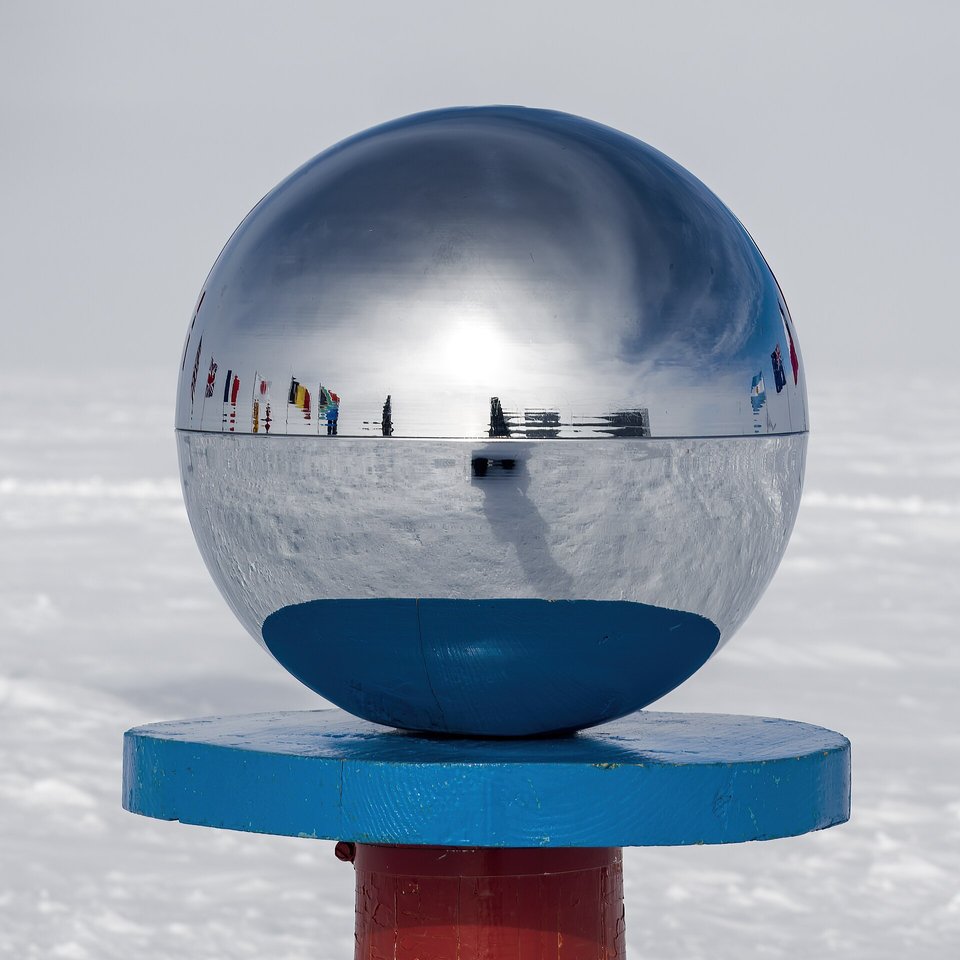 A large mirrored sphere on a blue disc above a red post, its surface reflecting a line of flags, with pale snow and sky behind
