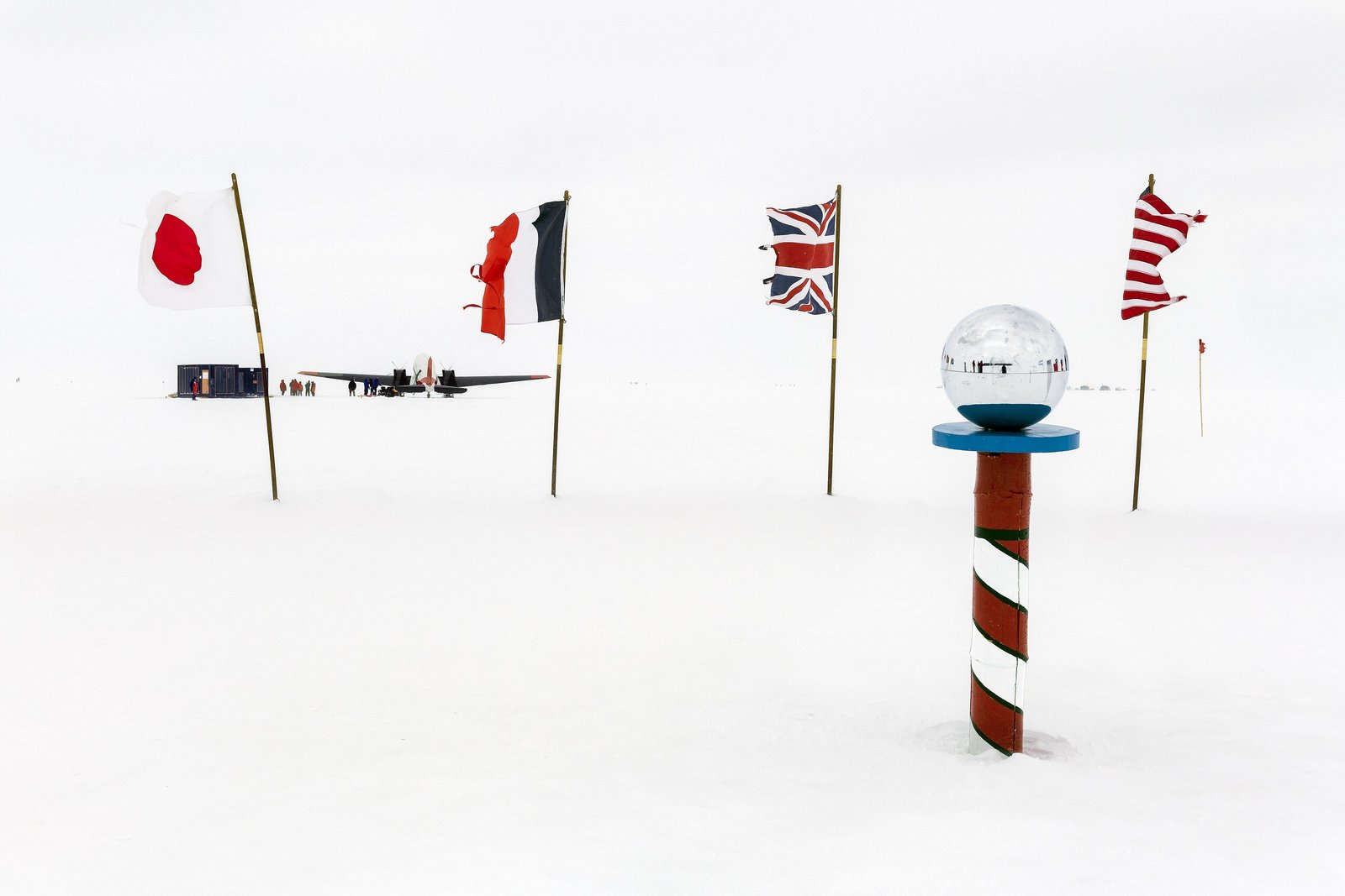 A ring of national flags and a short red-and-white striped pole topped by a mirrored ball on flat white snow under a white sky, a small aircraft and a huddle of tiny figures in the distance